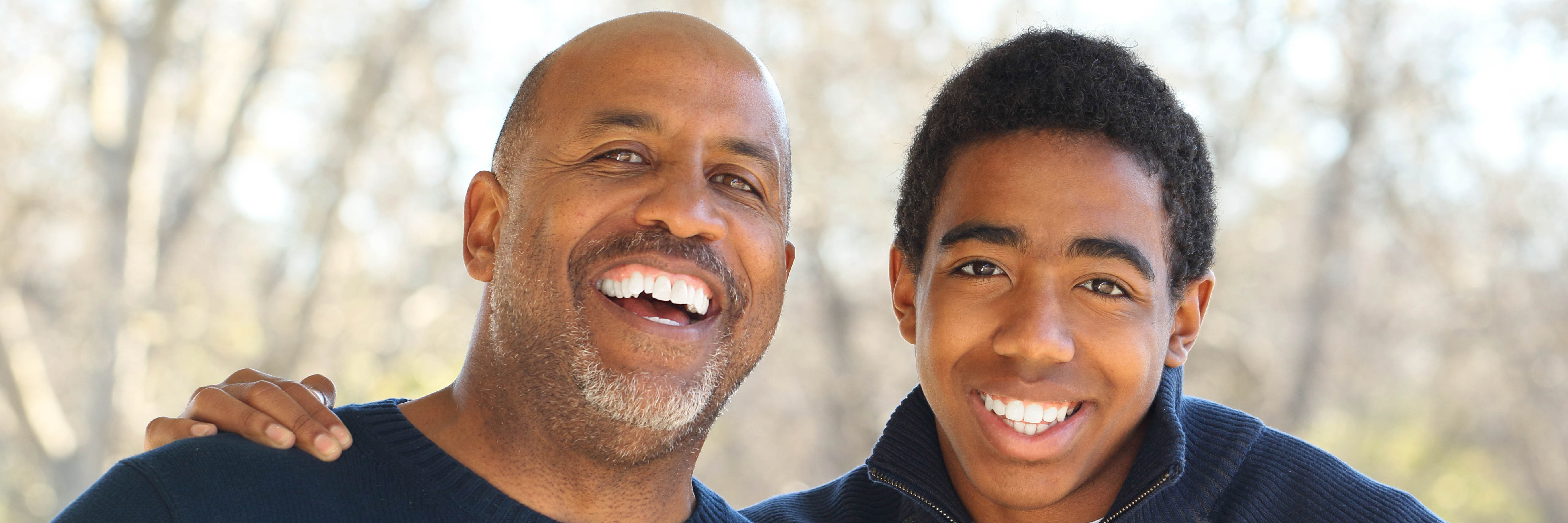 A father and his teenage son, side-by-side, smiling outdoors. 